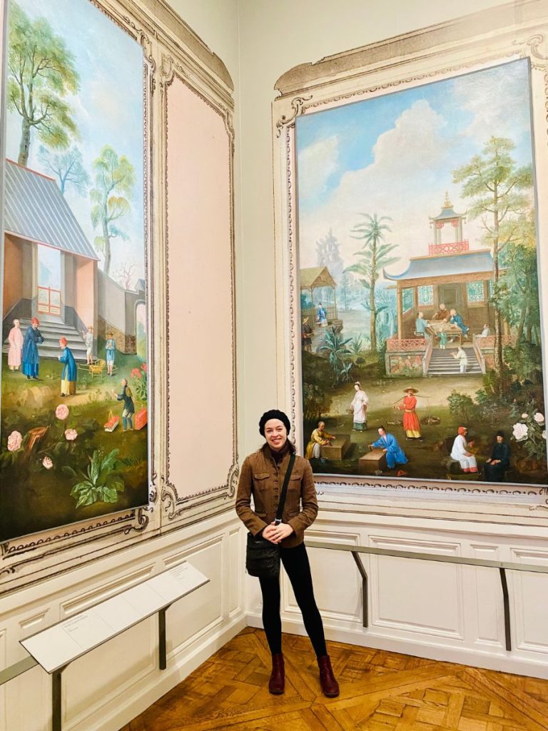 Agni posing in the Palace of Versailles in front of large paintings showcasing Asian-style buildings, blending Eastern and Western artistic influences.