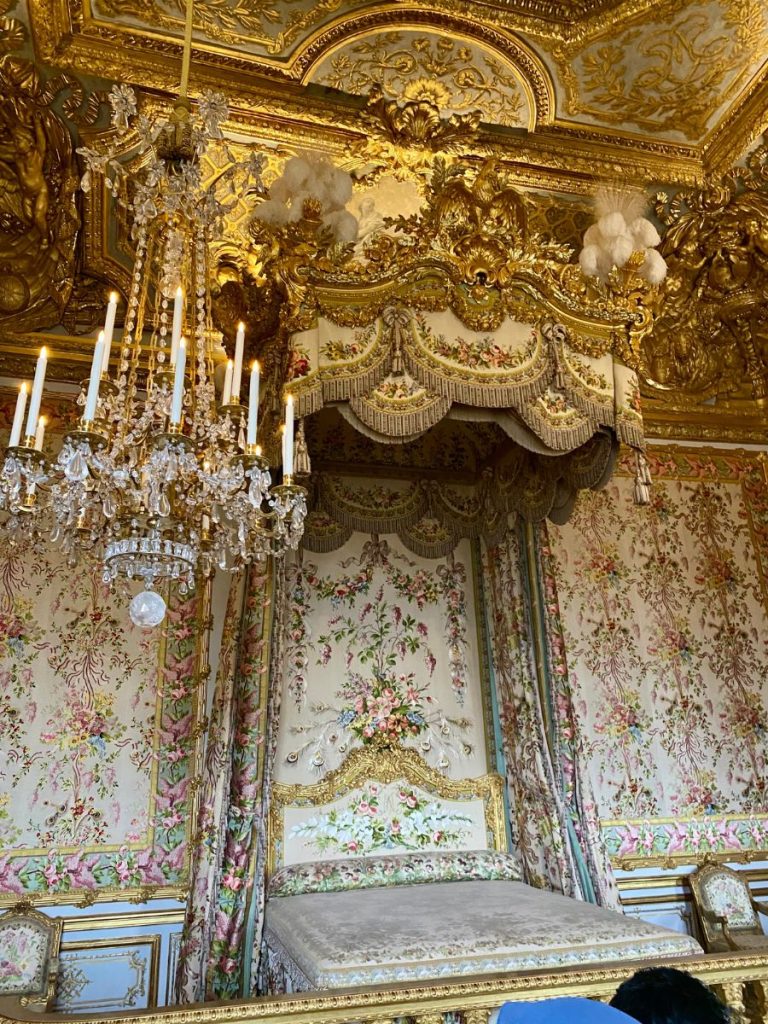 Lavish royal bed and glittering chandelier in a Palace of Versailles bedroom — one of the extravagant interiors that answers: is Versailles worth visiting?